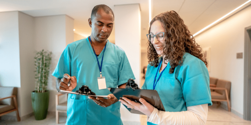 Two happy nurses in hallway, one holding clipboard, discussing patient care.